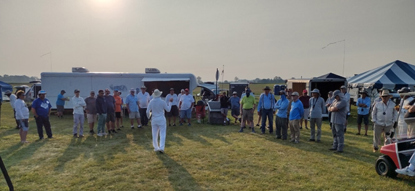 Large group of people gathered outdoors on grass, trailers in background.