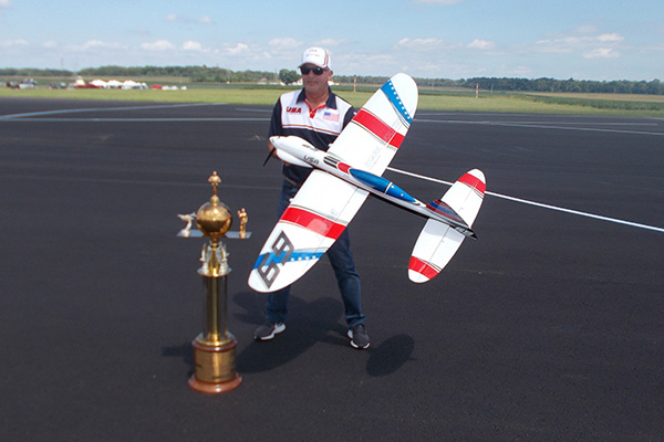 Man holding a model airplane next to a large trophy on an airfield runway.
