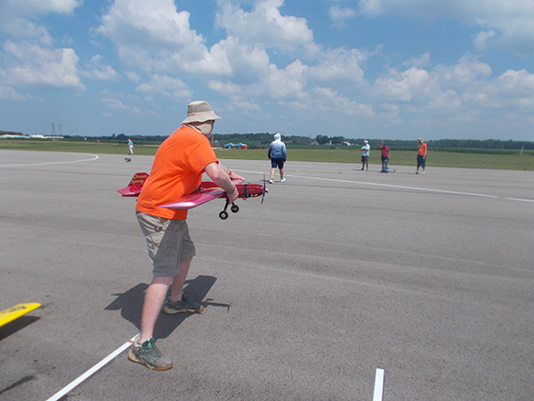 Man in hat launching model plane on runway under blue sky.