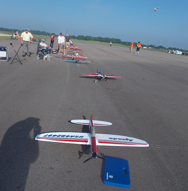Rows of model airplanes on a sunny tarmac, a small crowd observing nearby.