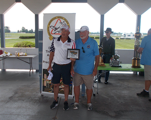 Two men in sports attire holding a framed award, standing in a bright pavilion.