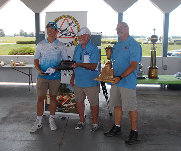 Three men holding trophies at an outdoor event, smiling.