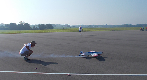 Toy plane on pavement with person crouching nearby; vast open sky and field in background.