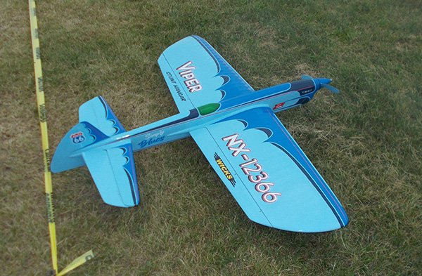 Blue and white model airplane on grass with a yellow measuring tape beside it.