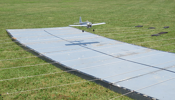 Small white model airplane landing on a large solar panel in a grassy field.