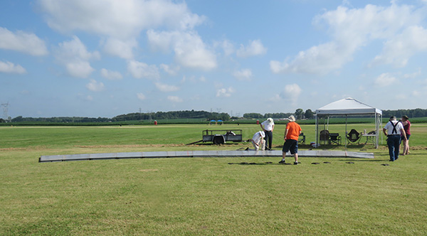People assembling a long metal structure on a grassy field under a blue sky.