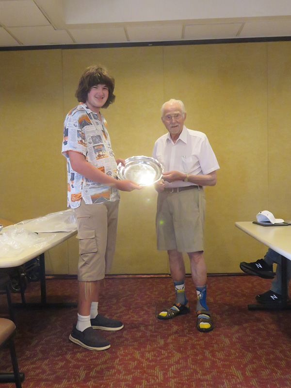 Young person and older adult holding a shiny award in a room with carpet and chairs.