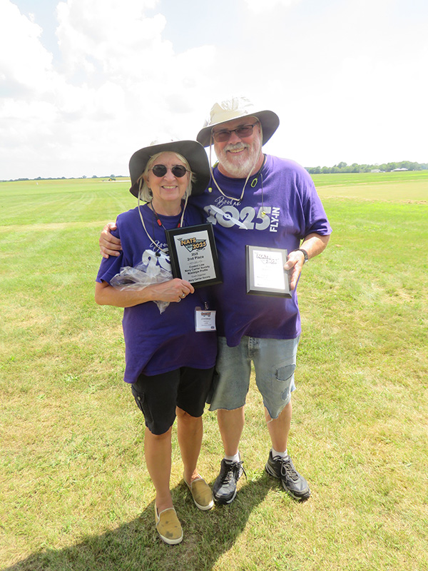 Two people in purple shirts and hats, smiling and holding plaques on a grassy field.