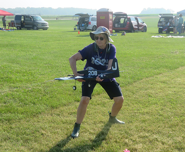 Person holding a model airplane on a grassy field, wearing a hat and sunglasses.