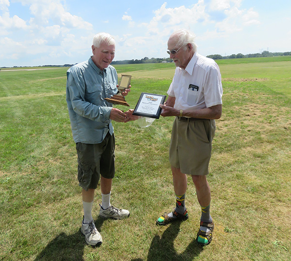 Two elderly men exchange awards outdoors on a grassy field.