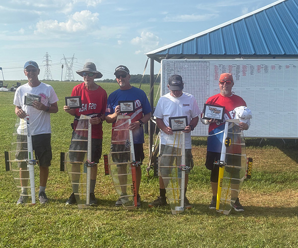 Five people outdoors holding rocket models and plaques, standing on grass.