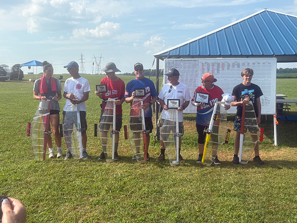 Seven people holding trophies, standing in a line outdoors on grass.
