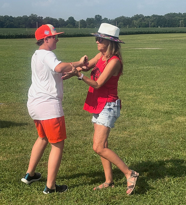 Two people wearing hats dance outdoors on a sunny grassy field.