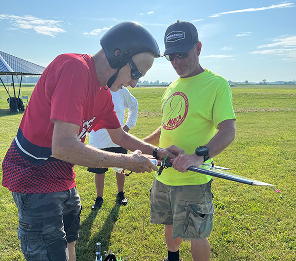 Two men with helmets prepping a small rocket outdoors on a sunny day.