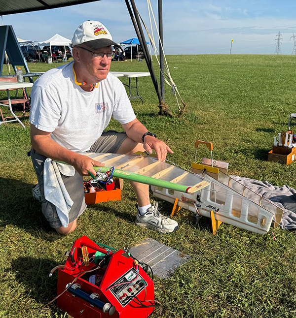 Man assembling a model airplane on grassy field, under clear sky.