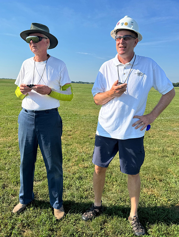 Two men in hats and sunglasses standing on grass with remote controls.