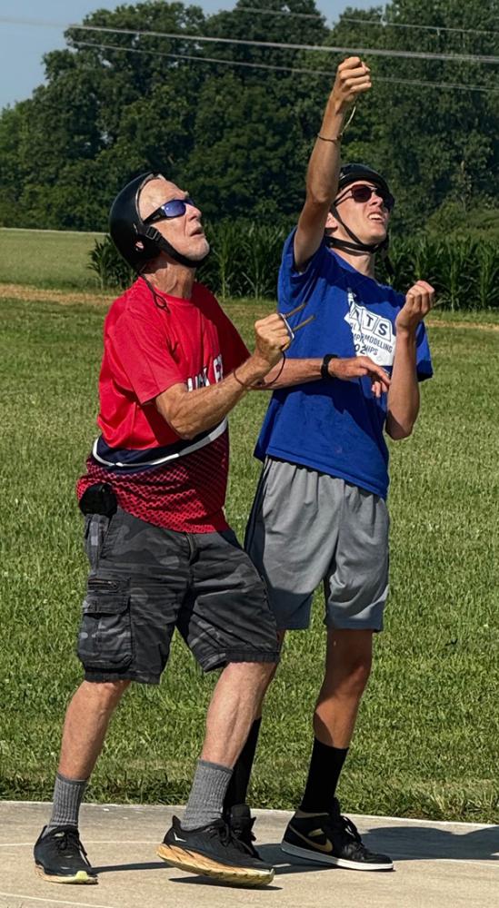 Two people flying kites in a grassy field, wearing helmets and sunglasses.