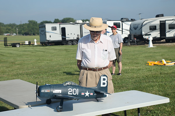Man observing model airplane on a table outside, RVs in the background.