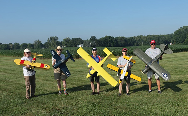 Five people hold large model airplanes on a grassy field.