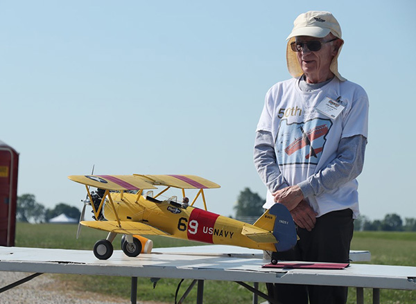 Man standing beside a model yellow biplane on a table outdoors.