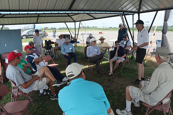 Group of people sitting under a canopy, some wearing hats and casual clothing.