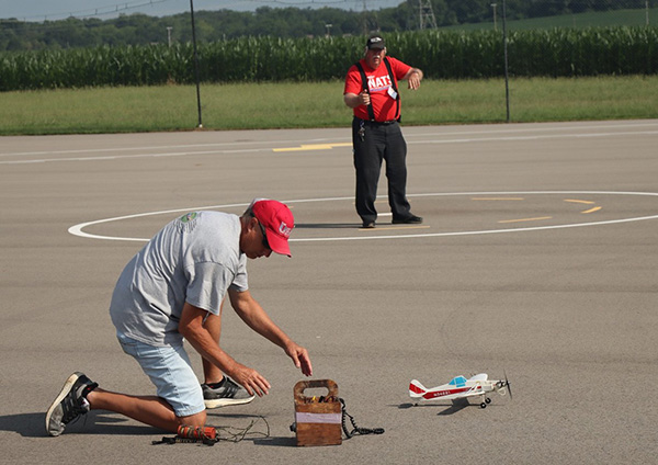 Man kneels on airfield with remote-controlled plane; another man stands in distance.