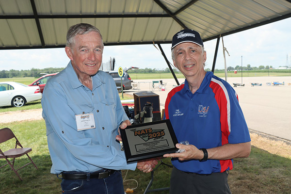 Two men in a pavilion holding a racing award plaque, smiling.
