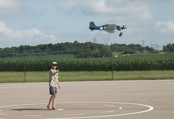 A small blue model airplane flying over a rural runway, with a person on the ground.
