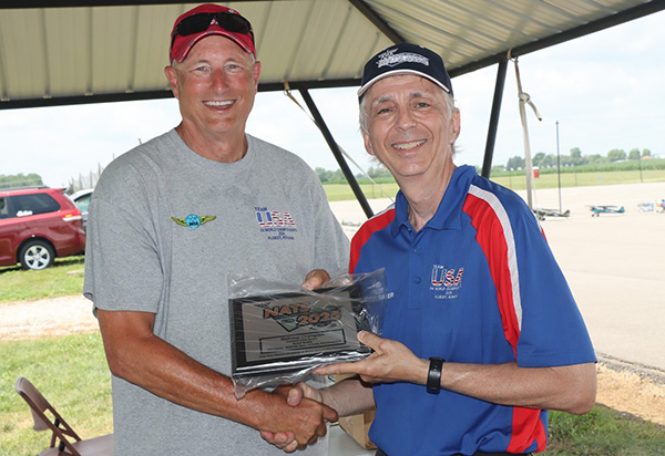 Two men smiling and shaking hands, one holding a plaque under a canopy at an outdoor event.
