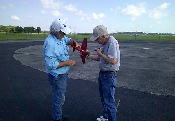 Two men examine a small red model airplane on a tarmac under a blue sky.