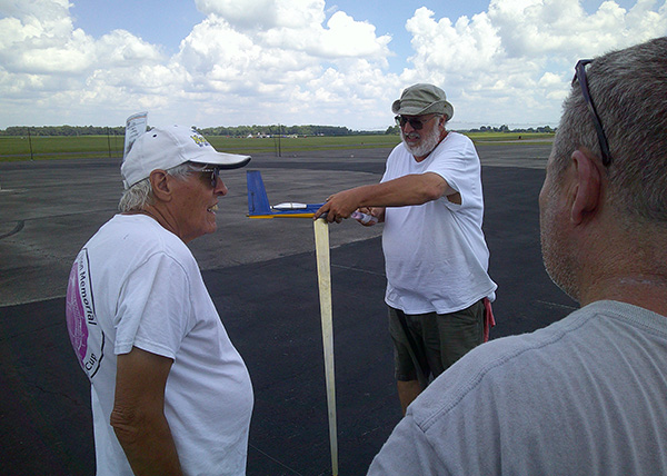 Three men in casual clothing, standing near a model airplane on an airfield.