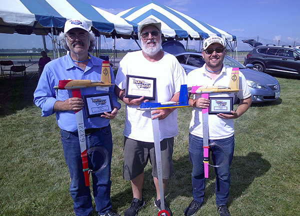 Three men holding model airplanes at an outdoor event with blue skies.