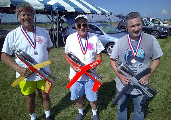 Three men holding model rockets, wearing medals, smiling outdoors.