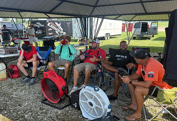 Four people sitting under a canopy, using fans to stay cool.