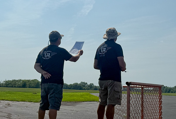 Two people in hats, outdoors, looking at a paper by a fence, under a clear sky.