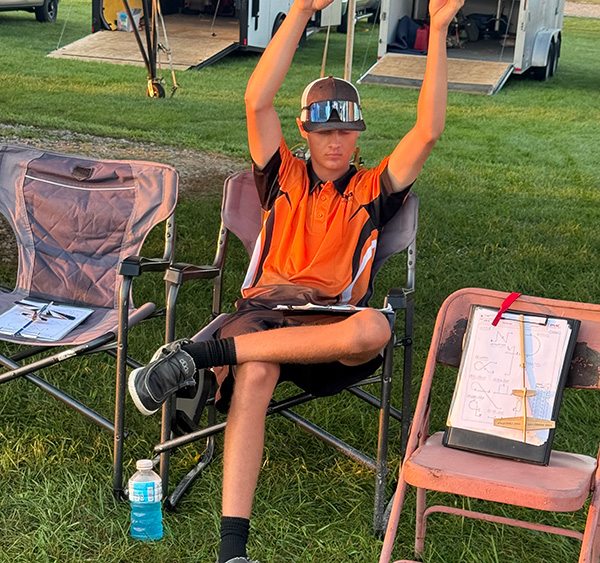 Boy in sunglasses and orange shirt sits in a folding chair, arms raised, outdoors.