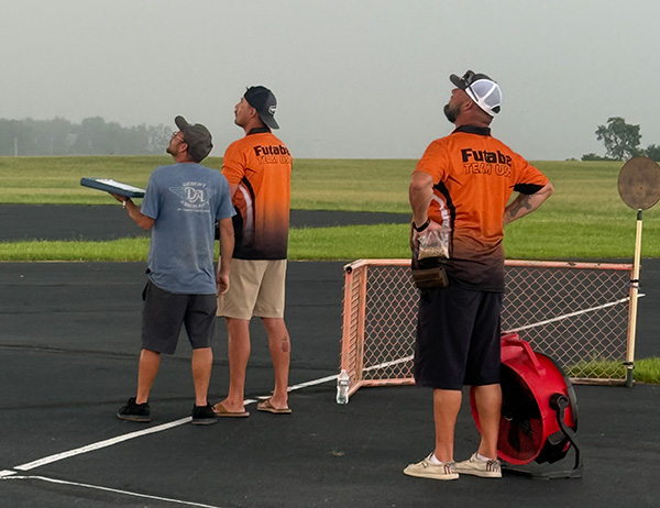 Three men observing a field, two wearing orange shirts, one holding a radio controller.