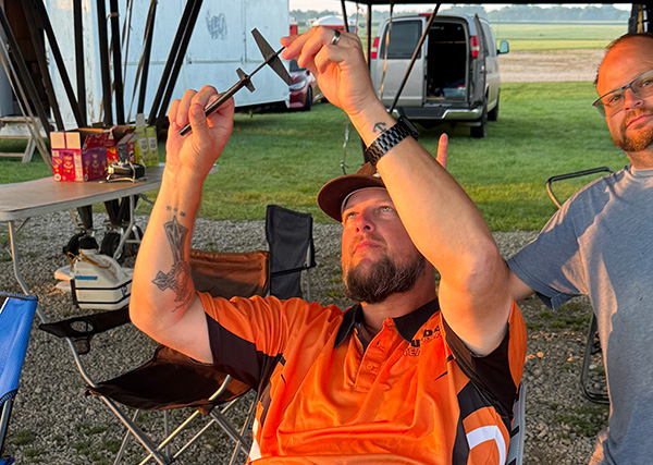 Man in orange shirt inspecting metal tool outdoors, another person in background.
