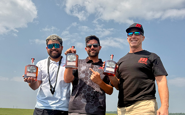 Three smiling men outdoors holding bottles, under a blue sky.