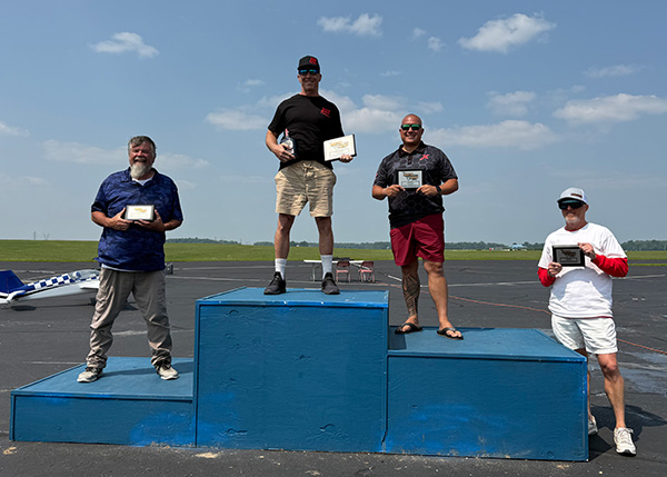Five men standing on winners' podium outdoors under a clear sky.
