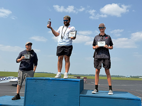Three men standing on a podium, holding certificates and a trophy, under a blue sky.