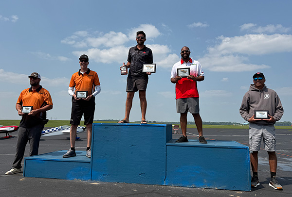 Five people on podiums holding awards outdoors, under a blue sky.