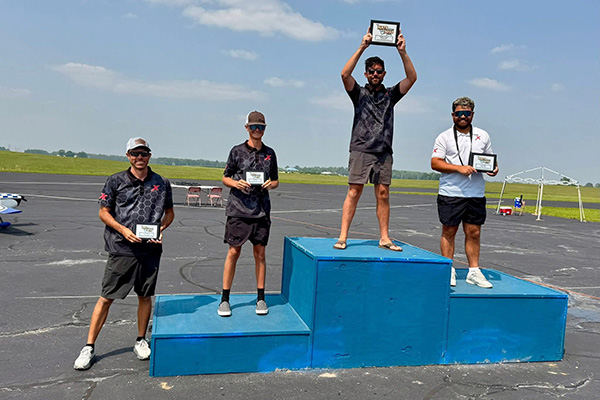 Four men holding plaques stand on a winners' podium outdoors, under a clear blue sky.