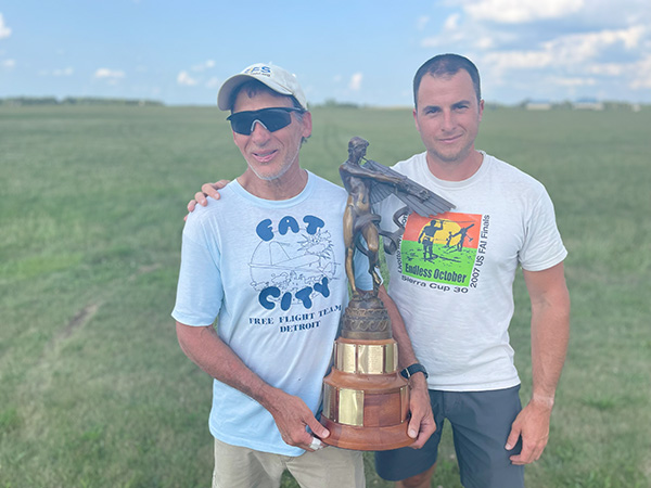 Two men standing on grass, holding a trophy, one wearing sunglasses.