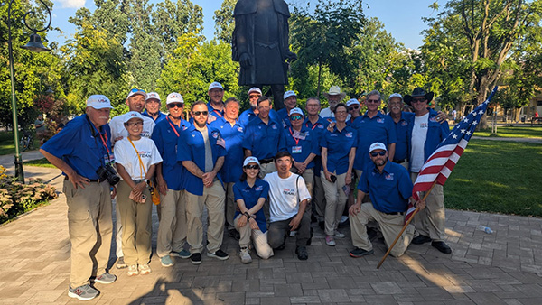 Group of people in blue shirts and white caps, some holding American flags, outdoors in a park.