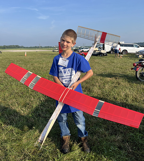 Boy holding a red model airplane in a grassy field.