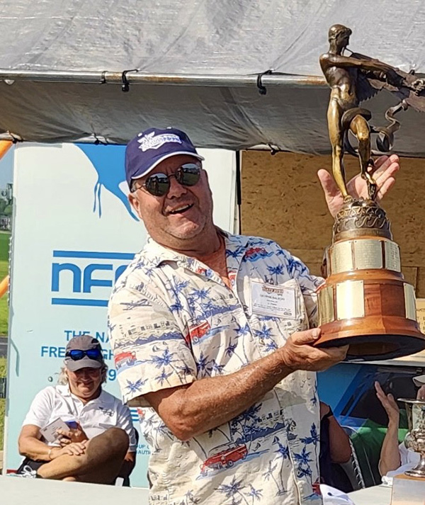 Man in a hat holding a large trophy, smiling under a canopy.