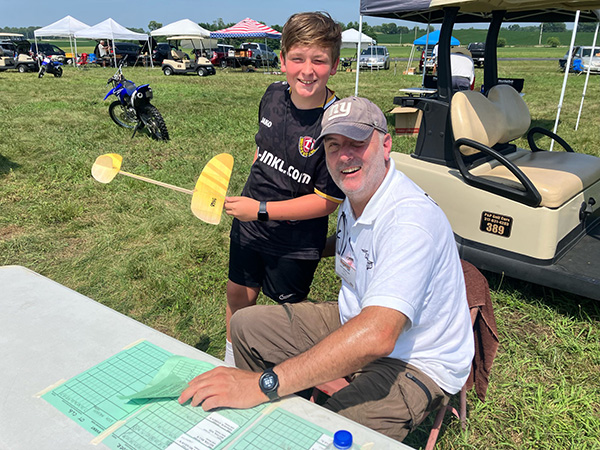 Boy holding model plane, smiling with seated man outdoors.
