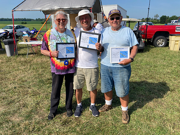 Three people smiling, holding framed certificates outdoors at a sunny event.