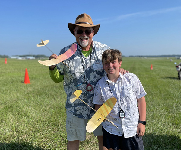 Man and boy holding model airplanes on a grassy field.